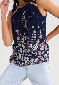 Marinblus utan ärmar i lätt, flödande tyg med blommönster i pastellfärger. Har plisseringar och en subtil metallisk kant vid halsringningen.