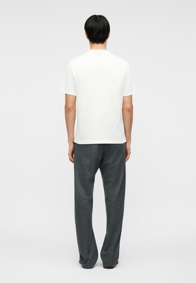 Άνδρας που στέκεται με την πλάτη στον θεατή, φοράει απλό λευκό κοντομάνικο πουκάμισο και σκουρόχρωμο γκρι, φαρδιά παντελόνια, σε ανοιχτό φόντο.