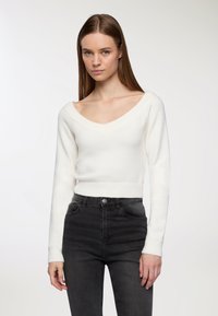 Maglione bianco lavorato a maglia con scollatura a V profonda, polsini e orlo a costine. Abbinato a jeans neri a vita alta. Texture liscia e design aderente.