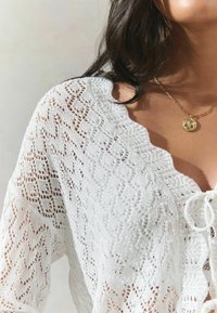 Haut en dentelle blanche avec un décolleté festonné et un détail de nœud. Présente des motifs ajourés complexes, une texture légère et une silhouette fluide.