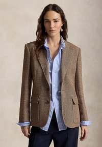 Femeie purtând un sacou maro cu model „herringbone” peste o cămașă bleu deschis cu nasturi și pantaloni închiși la culoare, stând pe un fundal bej simplu.