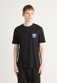 T-shirt noir à manches courtes en coton, avec une petite poche violette portant le logo "HUGO". Design simple, coupe décontractée, texture douce.