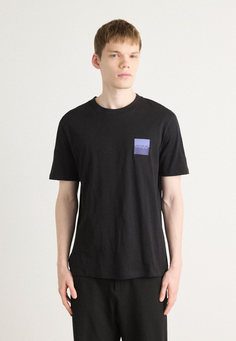 T-shirt noir à manches courtes en coton, avec une petite poche violette portant le logo "HUGO". Design simple, coupe décontractée, texture douce.
