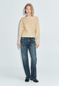 Donna in piedi con addosso un maglione beige a maniche lunghe, jeans scuri larghi e mocassini neri, contro uno sfondo chiaro uniforme.