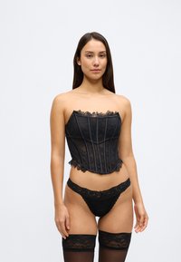 Fekete csipkés fűző függőleges varratokkal és csipkés szegéllyel, hozzá illő csipke tangával és combfixszel, amelyek hangsúlyozzák a részletek finomságát.