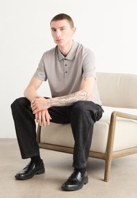 Grå polo-skjorte med to knapper, mørke jeans, sorte loafers, tatovert arm som hviler på knærne, sittende på en beige stol med treben.