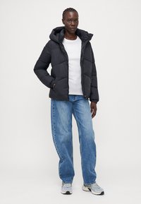 Svart puff jakke med hette, med diagonalt quiltede seksjoner og frontglidelås, båret over en hvit skjorte og lyseblå jeans.