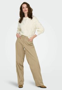 Femme aux cheveux bruns lâches portant un pull crème à motifs en losange, un pantalon beige large et des chaussures pointues brunes avec les mains dans les poches.