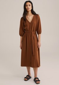 Brun, texturerad midi-klänning med djup V-ringning, ärm som slutar vid armbågen och puffärmar, samt ett bälte i tyg. Knappar fram och avslappnad passform. Svarta sandaler.