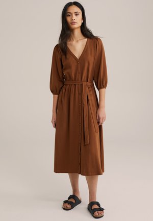 Robe midi marron texturée avec un décolleté en V profond, des manches bouffantes à longueur de coude et une ceinture en tissu. Devant à boutons et coupe décontractée. Sandales noires.