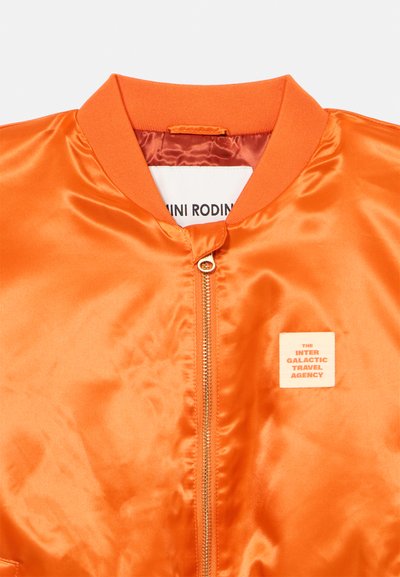 Πορτοκαλί σατέν μπουφάν βομβαρδισμού με ριμπ λαιμόκοψη, κεντρικό φερμουάρ και μικρό τετράγωνο μοτίβο που γράφει "Η Διαγαλαξιακή Ταξιδιωτική Υπηρεσία."