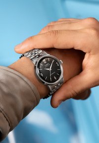 Silberne Armbanduhr mit schwarzem Zifferblatt, numerischen Stundenmarkierungen und einem Metallband. Die Hand ist am Handgelenk positioniert und zeigt die Uhr deutlich.