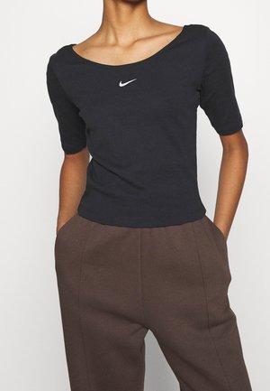 Czarny bawełniany top z krótkim rękawem, dekoltem w serek oraz białym logo Nike na piersi, zestawiony z brązowymi spodniami z wysokim stanem.