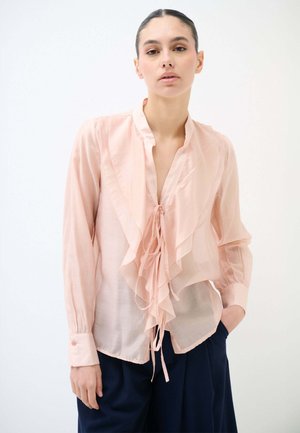 Donna che indossa una camicetta trasparente rosa pallido con dettagli arricciati e maniche lunghe, abbinata a pantaloni larghi blu scuro, in piedi davanti a uno sfondo bianco.