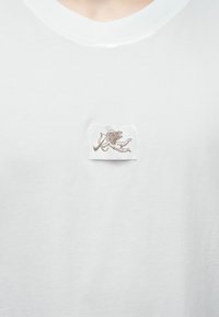 Fehér pamut póló klasszikus kerek nyakkivágással. Kis hímzett folt díszíti, amely egy íjjal és nyíllal rendelkező szárnyas alakot ábrázol.