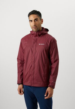 Bărbat îmbrăcat într-o jachetă Columbia bordo cu glugă și pantaloni albastru închis, stând în fața unui fundal simplu gri deschis.