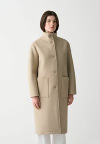 Beige ullkappa med hög krage, knäppning fram och två framfickor. Har rak skärning och mjuk textur.