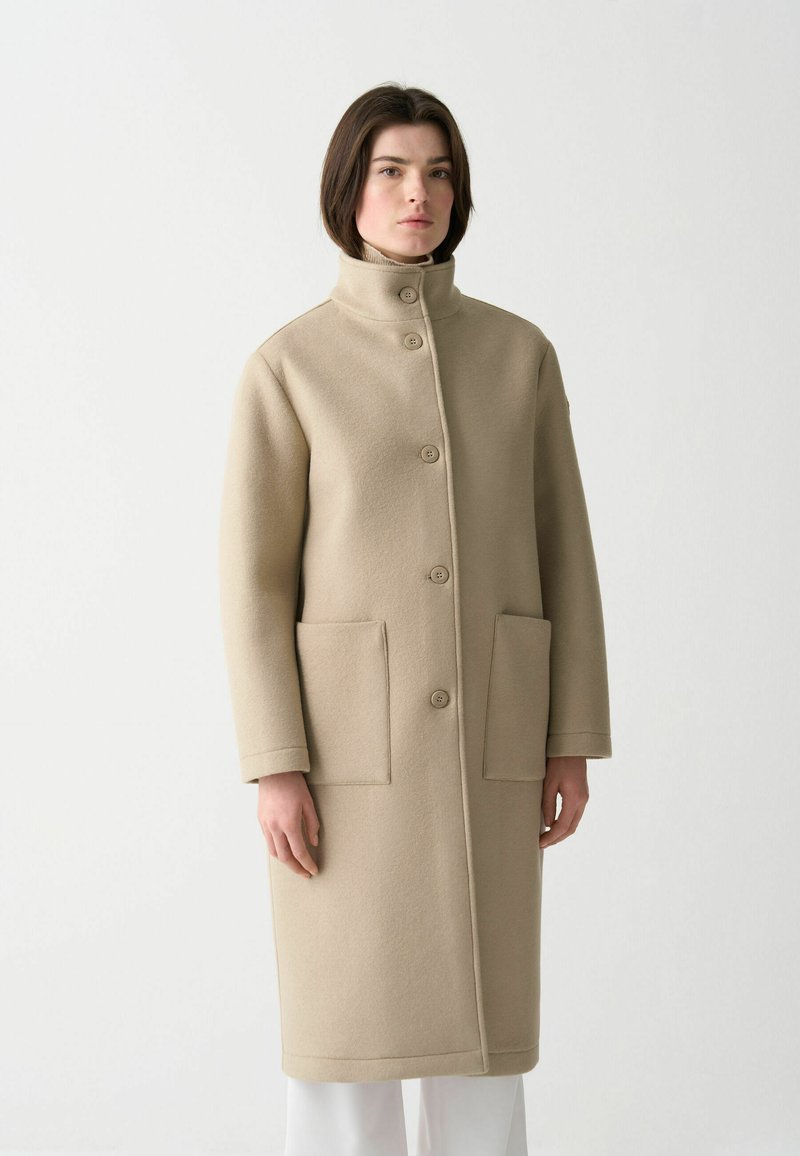 Beige ullkappa med hög krage, knäppning fram och två framfickor. Har rak skärning och mjuk textur.