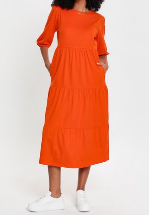 Orangenes Baumwollkleid mit kurzen Puffärmeln, gestuftem Design und Seitentaschen, kombiniert mit weißen Sneakers für einen lässigen Look.