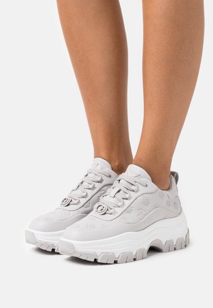 Fötter som bär ljusgrå klumpiga sneakers med prägling och tjocka vita sulor, stående mot en simpel vit bakgrund.