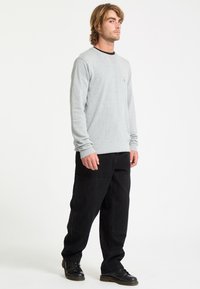 Jeune homme debout portant une chemise à manches longues gris clair, un pantalon noir et des chaussures noires sur un fond blanc uni.