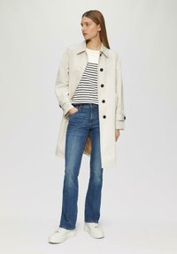 Hellbeige Trenchcoat mit runden Knöpfen, gestreiftem Hemd, dunkelblauen Jeans und weißen Sneakers. Besitzt einen lockeren Schnitt und lange Ärmel.
