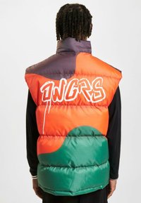 Farverig puffer vest med sektioner i orange, grøn og lilla. Bemærkelsesværdig hvid graffiti-tekst og quiltet tekstur med en høj krave.
