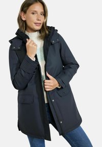 Veste imperméable bleu marine foncé avec capuche, fermeture éclair à l'avant, boutons-pression et deux poches avant. Texture douce et lisse, design décontracté.