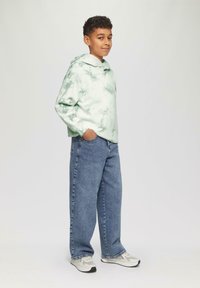 Zielona bluza z efektem tie-dye, z długimi rękawami, lu�źnym krojem i kieszenią kangurką, połączona z luźnymi niebieskimi dżinsami i jasnymi trampkami.