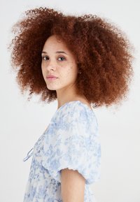 Vestido floral azul claro com mangas bufantes e textura fluida. A modelo tem cabelo encaracolado castanho-avermelhado e sardas, e está de pé contra um fundo simples.