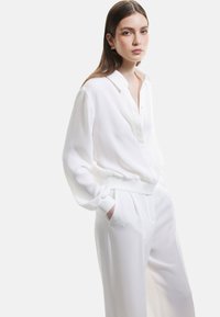 Blusa bianca leggera con chiusura a bottoni sulla parte anteriore e polsini a coste, abbinata a pantaloni bianchi su misura con pieghe e tasche laterali.