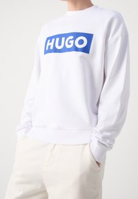 Biały sweter z odważnym niebieskim prostokątnym graficznym wzorem z napisem "HUGO" w białym kolorze. Projekt posiada ściągacze na mankietach i okrągły dekolt.