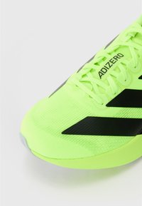 Zapatilla deportiva verde neón con parte superior de punto texturizado y acentos negros, con punta redonda y suela plana, marcada con "ADIZERO."