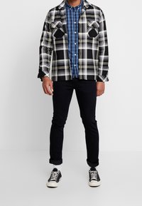 Chemise en flanelle à carreaux noir et blanc avec des accents jaunes portée sur une chemise à carreaux bleus boutonnée, associée à un jean foncé et des baskets noires.