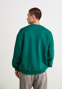 Green sweatshirt with a crew neck and dropped shoulders, featuring a textured fabric. The back has a contrasting pink accent near the neckline.