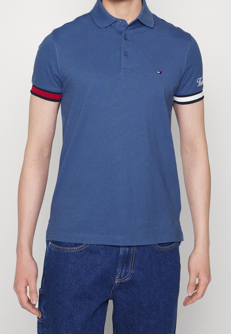 Navyblå polotröja i bomull med kort ärm, röd och vit detaljer, två knappar vid kragen och en liten logotyp på bröstet.