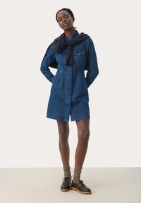 Denim shirt dress, light blue, with two chest pockets, button-down front, short sleeves, and a relaxed fit. Worn with navy sweater and brown shoes.