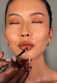 Påføring av makeup på leppene til en modell med en pensel. Modellen har en matt brun lepskygge, glatt hud og en nøytral bakgrunn.
