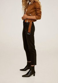Brązowa kurtka z sztucznej skóry z zamkiem błyskawicznym, dopasowany krój. Czarne jeansy z wysokim stanem z paskiem na klamrę, połączone z czarnymi botkami na kostkę.