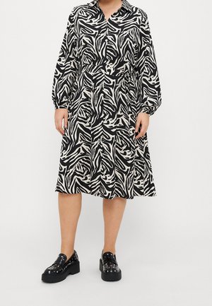 Kvinde iført en sort-hvid zebra-print langærmet kjole kombineret med sorte platform-pyntede loafers, stående mod en ensfarvet baggrund.