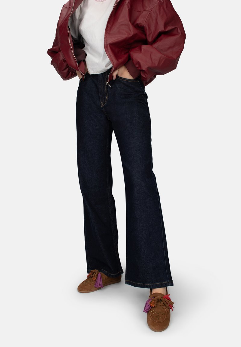 Persona vestida con jeans de pierna ancha azul oscuro, zapatos de ante marrón con borlas coloridas, una camiseta blanca y una chaqueta bomber roja, con las manos en los bolsillos.