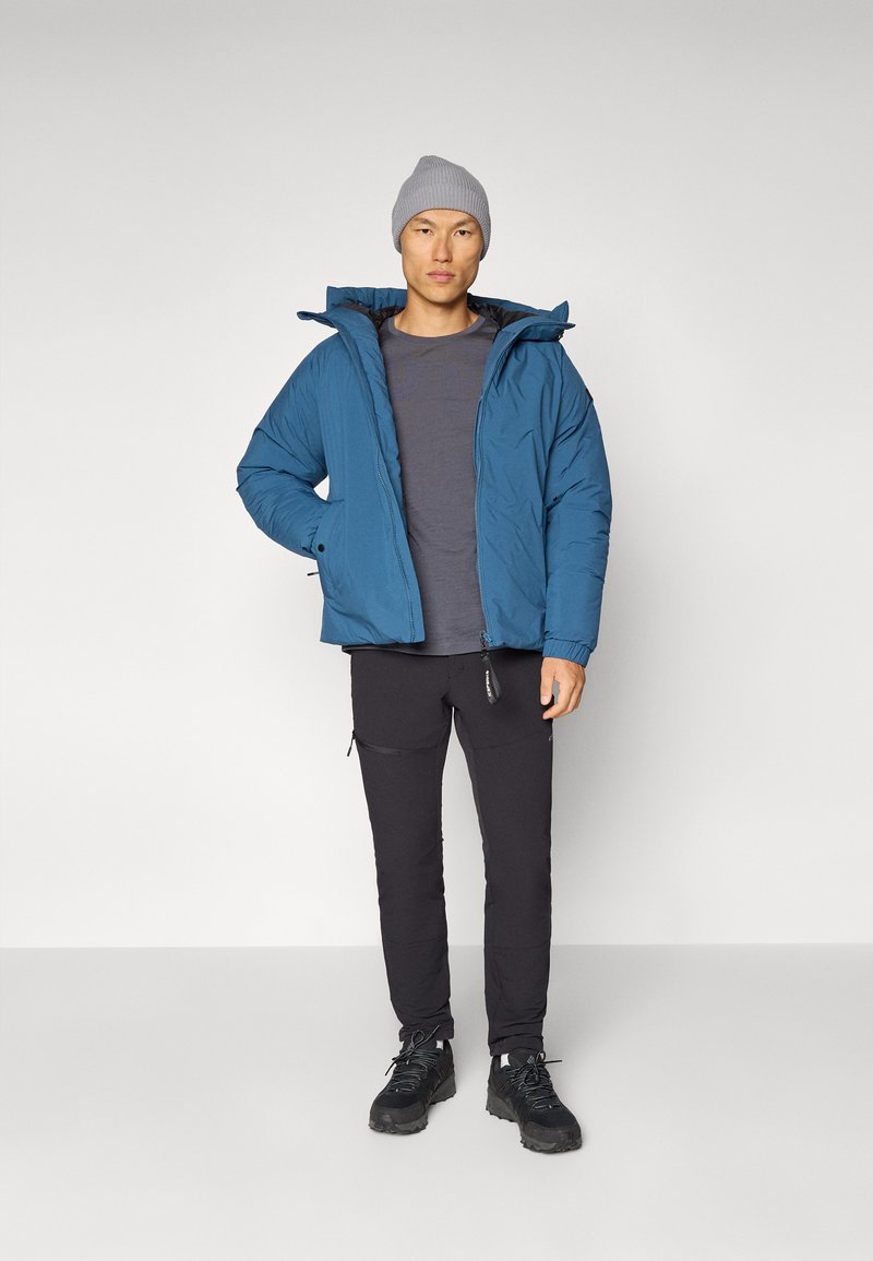 Blauer Pufferjacke mit Kapuze, getragen über einem grauen T-Shirt. Schwarze, schmal geschnittene Hose mit seitlichen Reißverschlüssen, kombiniert mit schwarzen Stiefeln und einer grauen Mütze.