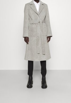 Persona che indossa un cappotto lungo grigio chiaro con cintura sopra pantaloni neri e stivaletti neri contro uno sfondo chiaro neutro.