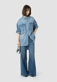 Chemise en denim avec deux poches poitrine, coupe loose ; associée à un pantalon en denim à jambe large. Le modèle tient des accessoires argentés brillants. Couleur bleu clair.