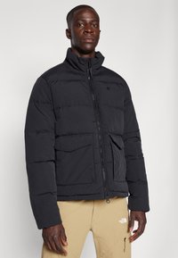 Doudoune noire avec un col haut, fermeture éclair à l'avant, deux grandes poches plaquées et un design matelassé, fabriquée en tissu lisse et mat.