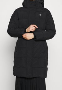 Černý péřový kabát s kapucí, s horizontálním prošíváním, zapínáním na zip, dvěma bočními kapsami a malým logem na hrudi.