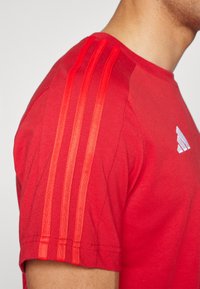 Rød treningst-skjorte laget av stoff, med tre vertikale tynne striper på skulderen og en liten hvit logo på brystet.