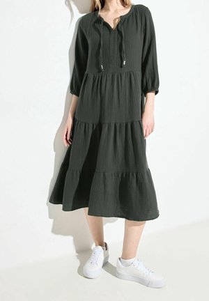 Kobieta w ciemnozielonej, warstwowej sukience midi z rękawami do łokci i białych trampkach, stojąca na białym tle.