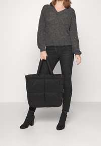 Bolso tote acolchado negro con diseño de acolchado; las asas son resistentes; expuesto con un suéter texturizado oscuro y jeans negros ajustados.