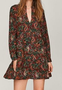Langärmliges Kleid in Dunkelgrün mit einem Paisleymuster in Rot, Weiß und Gelb. Verfügt über einen tiefen V-Ausschnitt und einen gestuften Rock.